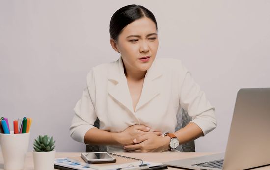 Isang babaeng nakaupo sa desk at nakahawak sa kanyang tiyan dahil sa sakit o masamang pakiramdam. 