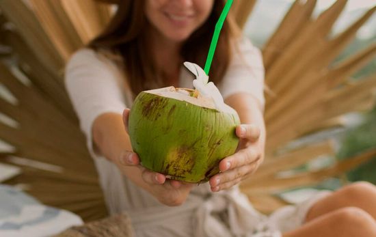 A person holding a fresh green coconut with a straw, ready to drink.