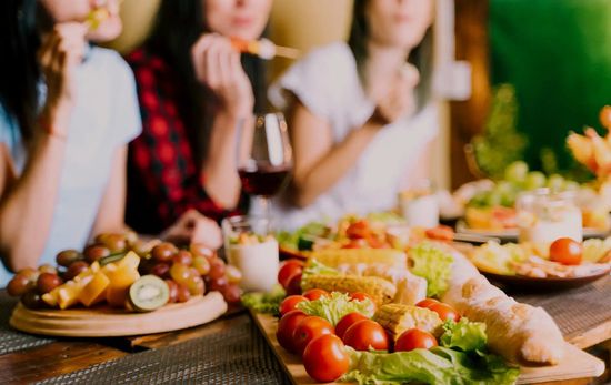 Different Healthy Food On A Table With Three Women Eating In The Background